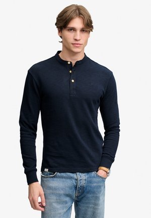 Ung mand iført en mørkeblå henley med lange ærmer og beige knapper samt lyseblå jeans, stående med den ene hånd i lommen.