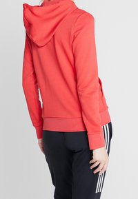 Sweatshirt corail avec capuche, doté de manches longues et d'un ourlet côtelé, porté avec un pantalon bleu marine qui présente trois bandes blanches sur le côté.