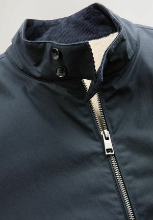 Navy jacket with a mock collar, featuring two black buttons and a silver zip closure. Inside lined with soft, cream-colored fabric.