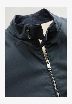 Marineblaues Jacket mit Stehkragen, ausgestattet mit zwei schwarzen Knöpfen und einem silbernen Reißverschluss. Innen mit weichem, cremefarbenem Stoff gefüttert.