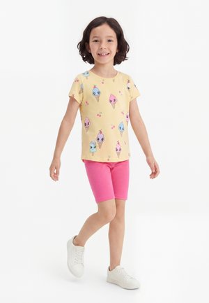 Ung pige går, iført gul t-shirt med farverige is og kirsebær print, lyserøde shorts og hvide sneakers på hvid baggrund.
