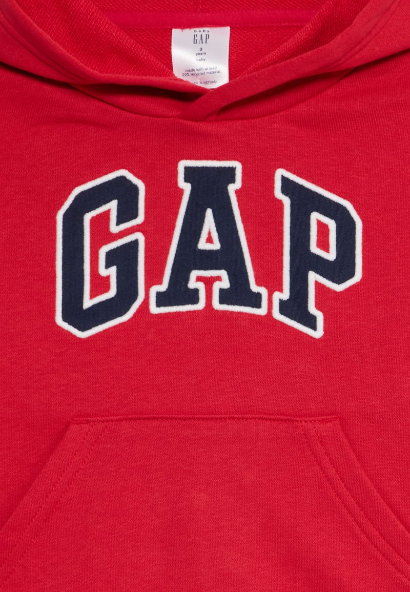 Sweat à capuche rouge avec un grand logo "GAP" en bleu marine cousu en blanc. Il possède une poche avant et un cordon de serrage pour la capuche. Texture de tissu douce.
