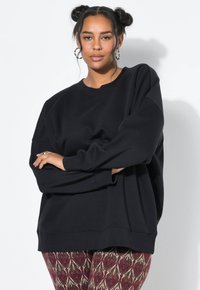 Sudadera negra de gran tamaño con cuello redondo y mangas largas, combinada con leggings rojos y dorados de patrón con diseños intrincados.