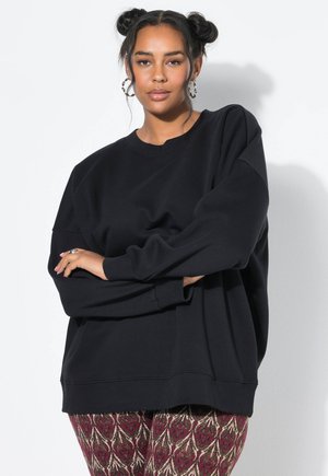 Mustat ylisuuria huppari, jossa on pyöreä kaula-aukko ja pitkät hihat, yhdistettynä kuvioituihin punaisiin ja kultaisiin leggings-housuihin, joissa on monimutkaisia kuvioita.
