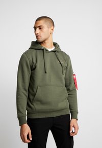 Olivengrønn hettegenser laget av myk stoff, med en frontlomme, snorhette og et lite emblem på brystet. Rød etikett på ermet.