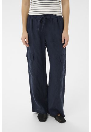 Person iført marineblå cargobukser med brede ben og bindebånd foran samt en hvid kortærmet, stribet skjorte, kombineret med sorte slip-on sko.