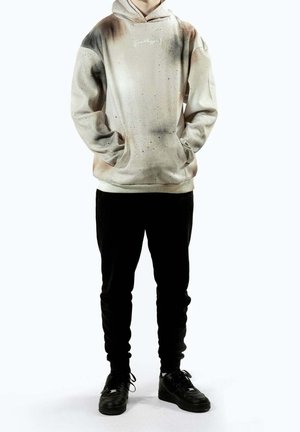 Hoodie met een zachte, gestructureerde crèmekleurige basis met spatten bruin en zwart, voorzakken in kangoeroestijl en een capuchon met trekkoord. Gecombineerd met een zwarte broek.