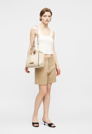 Frau trägt beige Shorts, weißes ärmelloses Oberteil, schwarze Sandaletten mit Absatz, hält eine cremefarbene Handtasche mit dem "Love Moschino"-Logo.