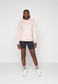 Camisola de algodão rosa claro com cordões, bolso frontal e logotipo branco, combinada com calções de bicicleta pretos e ténis brancos com meias riscas.