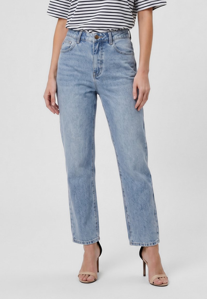 Femme portant un jean bleu clair droit taille haute, associé à des sandales beiges à talons hauts et un haut rayé noir et blanc.