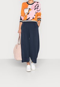 Dzianinowy sweter w kolorze pomarańczowym i różowym z czarnym wzorem kwiatowym; zestawiony z luźnymi granatowymi culottes i białymi trampkami. W zestawie również różowa torba.