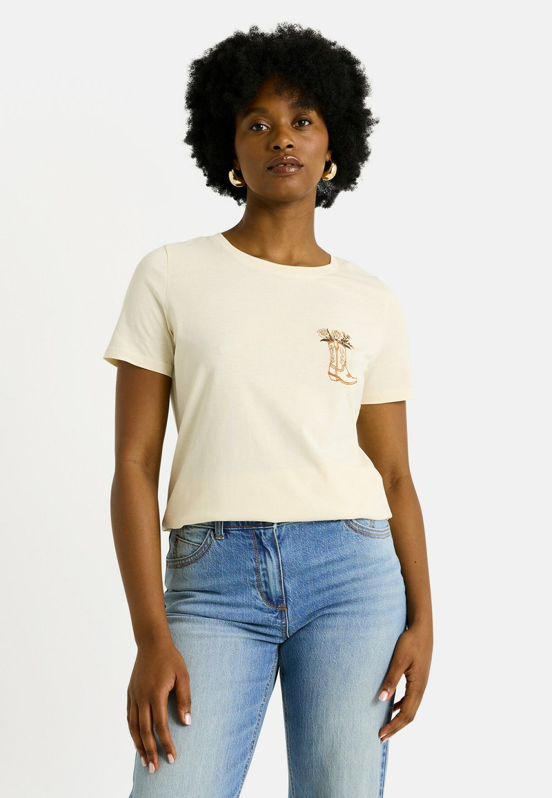 Camiseta de algodón color beige claro con mangas cortas, cuello redondo y un pequeño gráfico marrón de elementos florales dentro de botas en la parte izquierda del pecho.