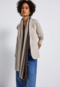 Beige kavaj med en figursydd design, i kombination med en texturerad grå och mörk scarf. Modellen bär en enkel topp och blå jeans.