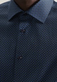 Mörkblå skjorta med små geometriska mönster av rutor och cirklar, knäppt, närbild på krage och övre bröst.