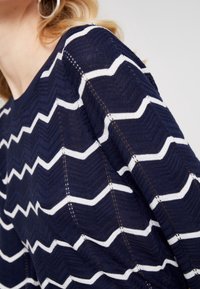 Pull bleu marine avec un motif en zigzag blanc, doté d'un tricot texturé et de détails en mailles ouvertes distincts le long des coutures.