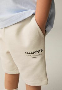 Pantaloni corti color crema realizzati in tessuto morbido, con tasche laterali e logo "ALLSAINTS" in nero. Una camicia azzurro chiaro sbuca dall'alto.