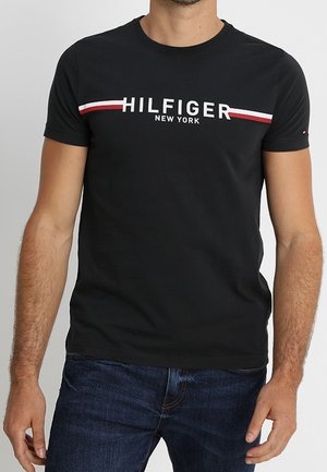 Mann trägt ein schwarzes T-Shirt mit kurzen Ärmeln mit der Aufschrift "HILFIGER NEW YORK" und roten und weißen Streifen über der Brust, kombiniert mit blauen Jeans.