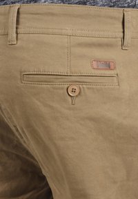 Beige canvasbukser med en baglomme med knaplukning og et lædermærke mærket "Solid." Glat tekstur, klassisk design.