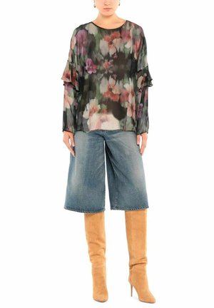 Donna che indossa una blusa floreale trasparente, pantaloni corti in denim larghi fino al ginocchio e stivali alti al ginocchio in suede color sabbia con tacco su uno sfondo bianco.