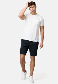 Hvid bomulds t-shirt med brystlomme og brun knap, parret med marineblå shorts og hvide sneakers, stående på en neutral baggrund.