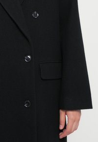 Cappotto di lana nero con una tasca sul petto, design doppiopetto con quattro grandi bottoni neri e una texture liscia.