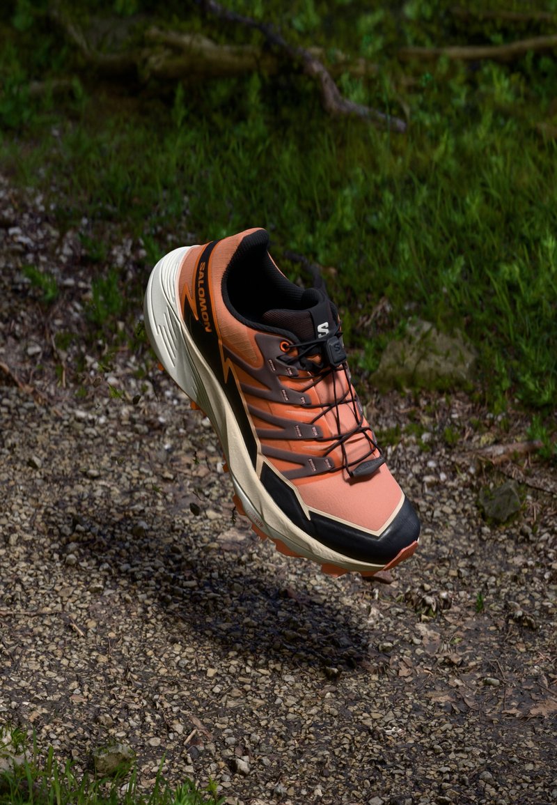 Běžecká obuv na trail s designem v oranžové, černé a béžové barvě. Obsahuje strukturovaný svršek, zesílenou špičku a podrážku s dobrým gripem.