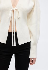 Λευκό ριπ πλεκτό πουλόβερ με μακριά μανίκια και κλείσιμο με δέσιμο μπροστά, συνδυασμένο με μαύρο παντελόνι με ψηλή μέση που αφήνει ακάλυπτη την κοιλιακή περιοχή.