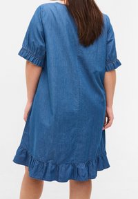 Vestido de mezclilla en azul medio con corte relajado, mangas cortas con volantes y un dobladillo en capa con volantes. Textura suave y diseño sencillo.
