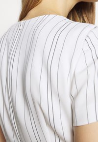 Femme aux cheveux châtain clair portant un vêtement blanc avec de fines rayures verticales noires et une fermeture éclair au dos au niveau du cou, vue de dos.