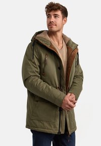 Parka verde con forro de felpa beige, acentos marrones, capucha ajustable y bolsillos con cremallera. Fabricada con tela duradera para brindar calidez.