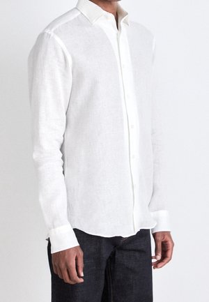 Homme portant une chemise blanche à manches longues boutonnée et un pantalon sombre, debout devant un fond clair uni.