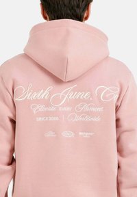 Sudadera rosa con un gran diseño de texto blanco en la parte posterior, que presenta "Sixth June, Co," y varios eslóganes. Tejido suave, ajuste casual.