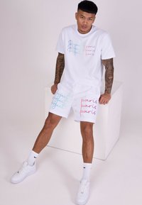 Camiseta blanca de algodón con el texto azul y rosa "Proyecto París". Los pantalones cortos blancos a juego presentan un texto similar en varios colores. Los brazos del actor están tatuados.
