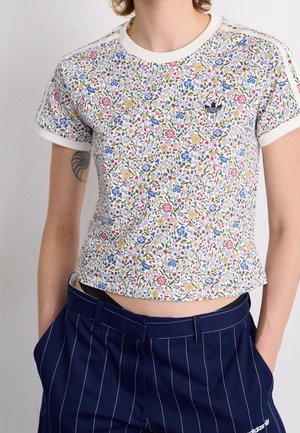 Personne portant un t-shirt court à imprimé floral avec le logo Adidas et un pantalon bleu marine à fines rayures, les mains dans les poches.
