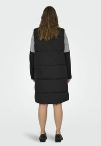 Svart pufferväst med en lång, vadderad design, som har grå och svarta tygpaneler på axlarna och långa ärmar.