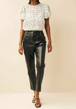 Femme portant un chemisier blanc à manches courtes avec des pois noirs, un pantalon en cuir noir taille haute et des sandales noires à bout ouvert.