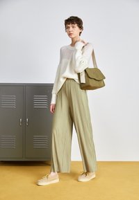 Suéter de punto crema, pantalones plisados de color verde claro y zapatos de plataforma beige. Bolso de hombro verde oliva. El fondo presenta taquillas grises.