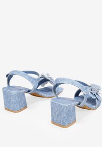 Sandalias de tacón block de denim con parte trasera abierta, correas de tobillo y borlas decorativas. Color azul claro con material de denim texturizado.