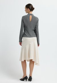 Grauer Strickoberteil mit hohem Kragen und einem Schlüssellochverschluss hinten, kombiniert mit einem fließenden, cremfarbenen asymmetrischen Rock und schwarzen High Heels.