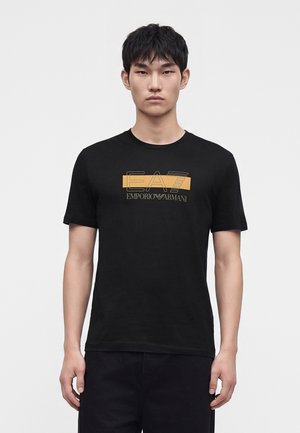 Jeune homme aux cheveux noirs courts, regardant devant, portant un t-shirt noir avec un logo Emporio Armani EA7 jaune et blanc au centre de la poitrine.