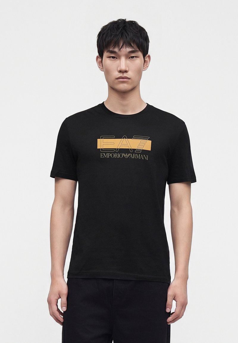 Junger Mann mit kurzen schwarzen Haaren, der nach vorne schaut und ein schwarzes T-Shirt mit gelbem und weißem Emporio Armani EA7-Logo auf der Brustmitte trägt.