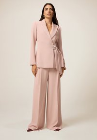 Blushkleurige blazer met een diepe V-hals, lange mouwen en een decoratieve broche; gecombineerd met wide-leg broeken in hetzelfde materiaal en kleur.
