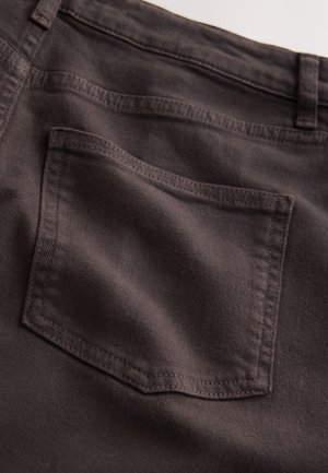Tumši brūnas džinsa bikses tuvplānā, ar vienu aizmugurējo kabatu ar pastiprinātu šuvumu, gludu tekstūru un klasisku piegriezumu.