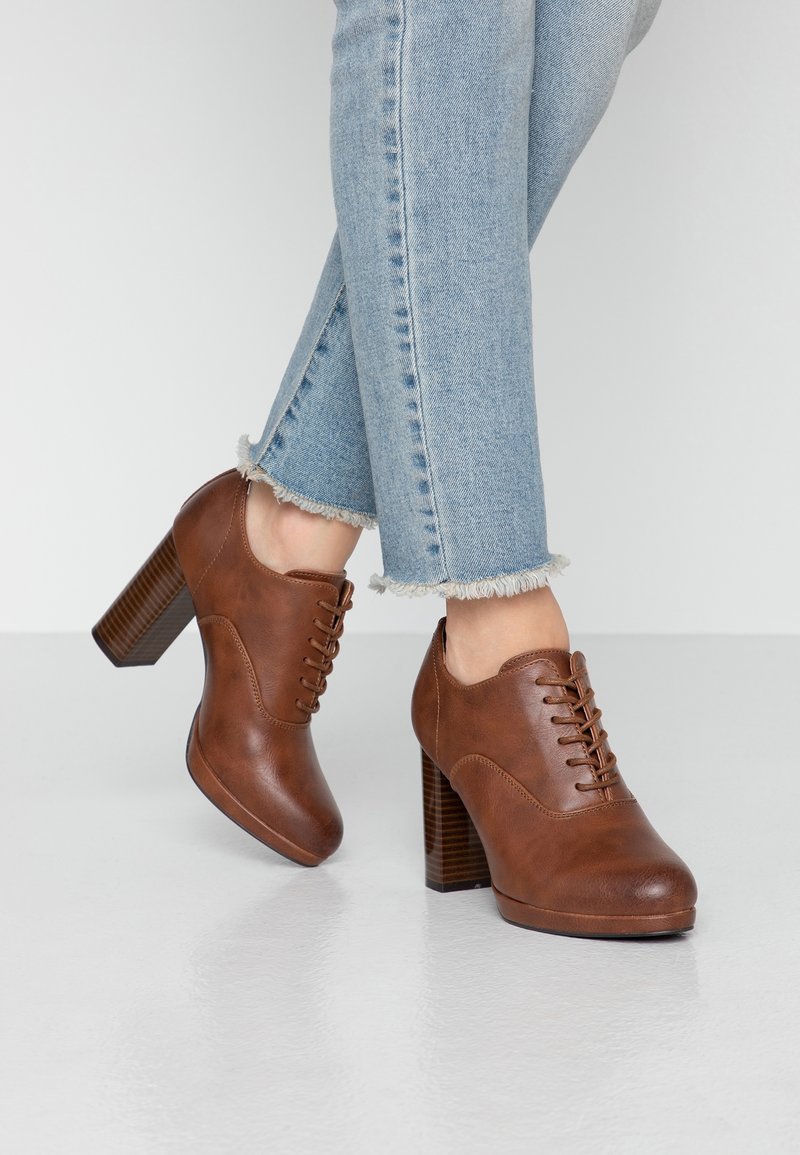 Chaussures à talons hauts en cuir marron à lacets portées avec un jean bleu clair à ourlet effiloché sur un sol gris clair et un fond blanc.
