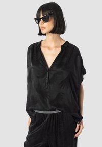 Črna bluzica s kratkimi rokavi in V-izrezom, ki ima subtilen ročni tisk. Narejena iz mehkega blaga, v kombinaciji z teksturiranimi črnimi hlačami.