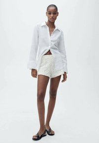 Witte linnen shirt met open kraag, gecombineerd met crèmekleurige kanten shorts. Zwarte sandalen maken de look compleet. Effen witte achtergrond.