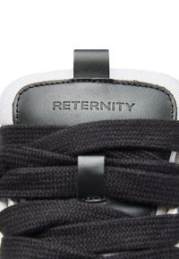 Czarne sznurowadła przeplatane przez teksturowane białe sportowe buty. Na górze znajduje się czarna skórzana metka z wytłoczonym napisem „RETERNITY”.