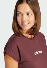 T-shirt bordeaux con maniche corte arrotolate, con il logo "adidas" bianco sul petto. Materiale in cotone morbido con una vestibilità comoda.