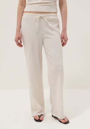 Person wearing loose, textured white drawstring pants and black square-toe sandals, standing against a plain light background.