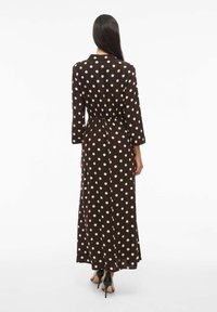 Vestido maxi de lunares marrones con cintura entallada, mangas largas y cuello, que presenta una textura de tela suave y un diseño de largo hasta el tobillo.